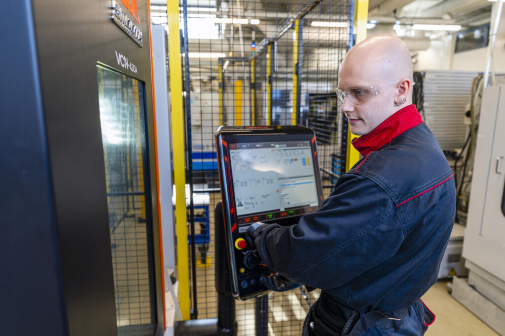 Henkilö seisoo teollisuuslaitteiston edessä ja käyttää kosketusnäytöllistä ohjauspaneelia. Hänellä on suojalasit ja tummansininen työasu punaisilla yksityiskohdilla.