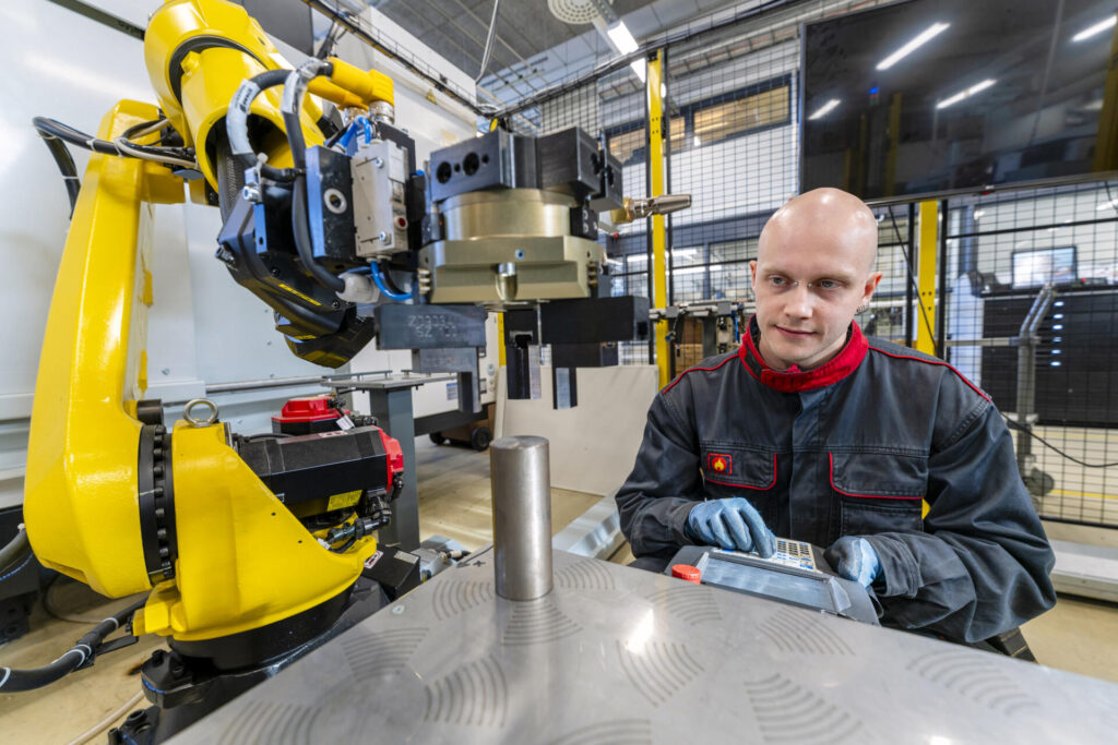 Mies työskentelee tehtaassa robotin vieressä, käyttäen ohjauslaitetta. Robotti on keltainen ja se liikuttaa metalliosaa.