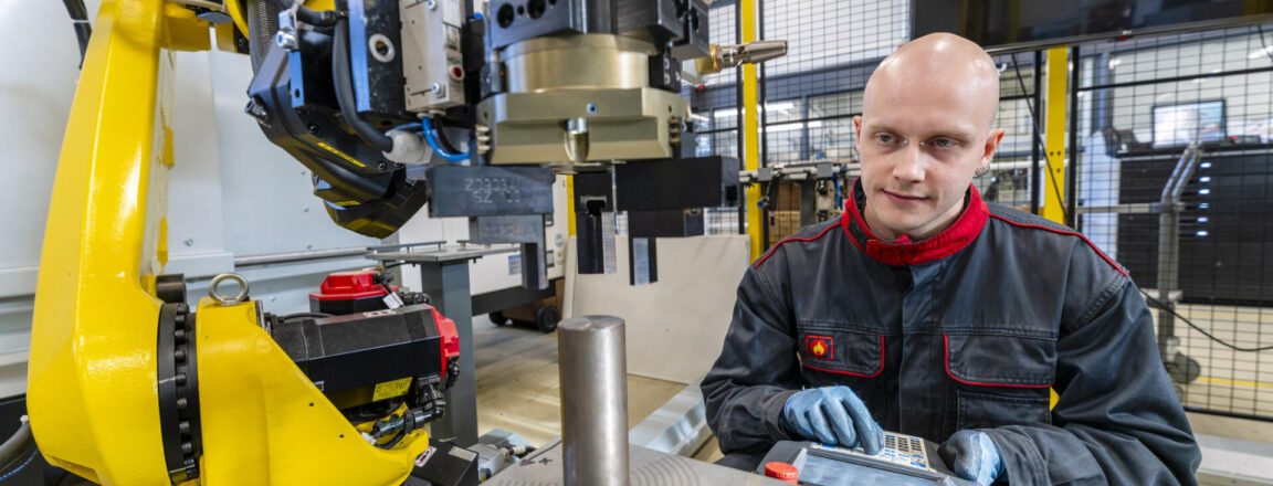 Mies työskentelee tehtaassa robotin vieressä, käyttäen ohjauslaitetta. Robotti on keltainen ja se liikuttaa metalliosaa.