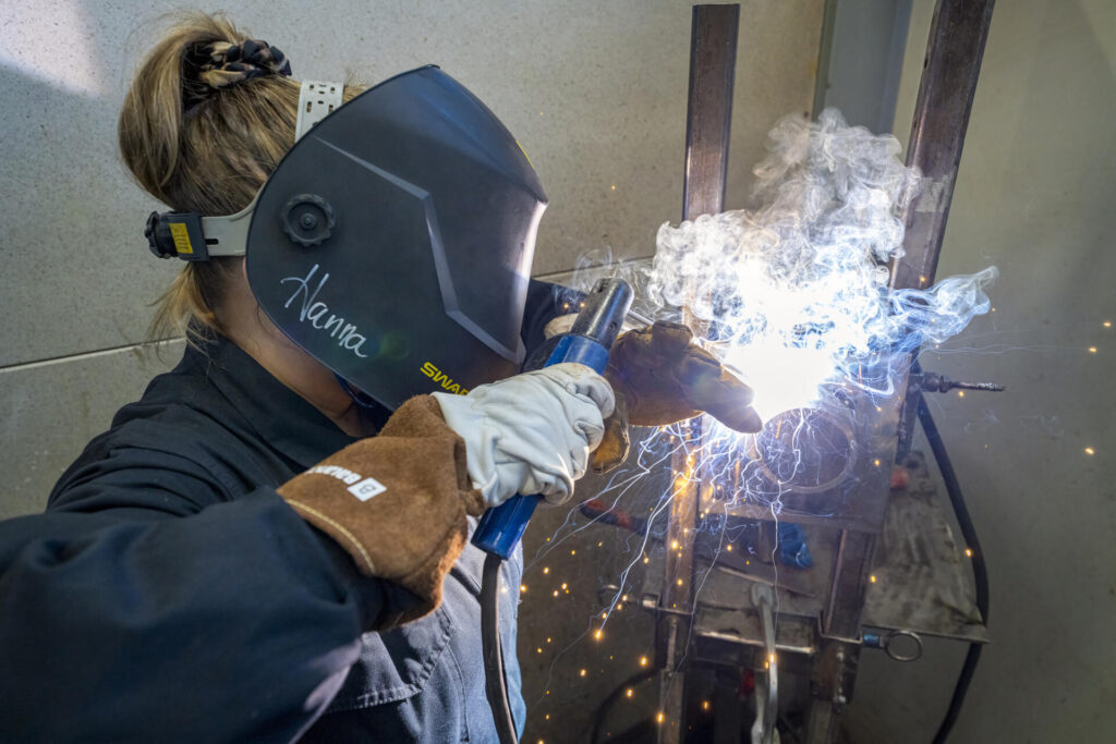 Henkilö hitsaamassa suojakypärä päässään. Kipinöitä lentää ympärillä, ja kirkas valo syntyy hitsausprosessista.