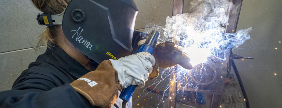 Henkilö hitsaamassa suojakypärä päässään. Kipinöitä lentää ympärillä, ja kirkas valo syntyy hitsausprosessista.