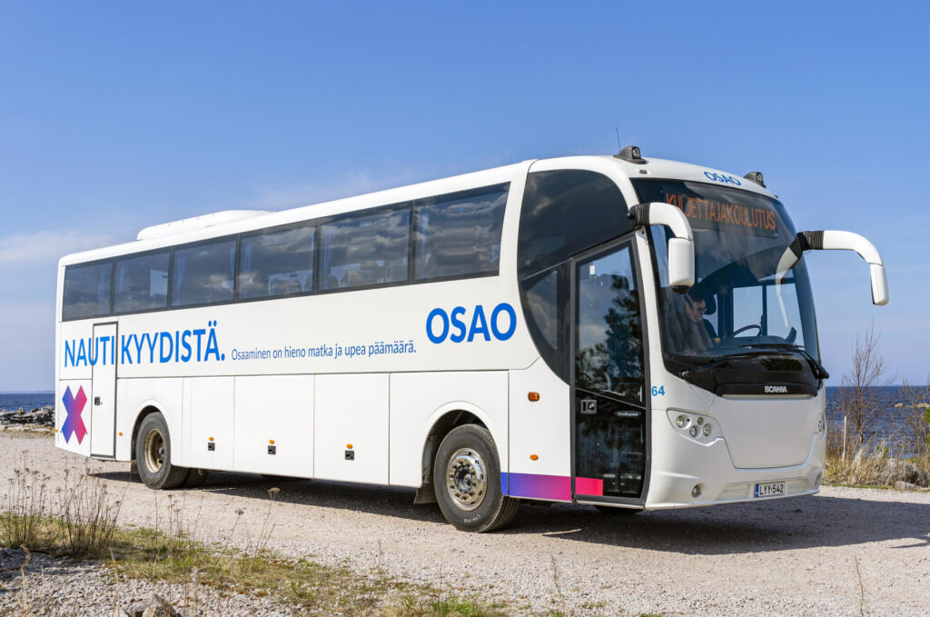Valokuva valkoisesta linja-autosta, jossa on siniset tekstit 'NAUTI KYYDISTÄ' ja 'OSAO' sivulla. Linja-auto on pysäköity soratielle meren rannalla, ja taustalla näkyy sininen taivas. Linja-auton edessä lukee 'KULJETTAJAKOULUTUS'.