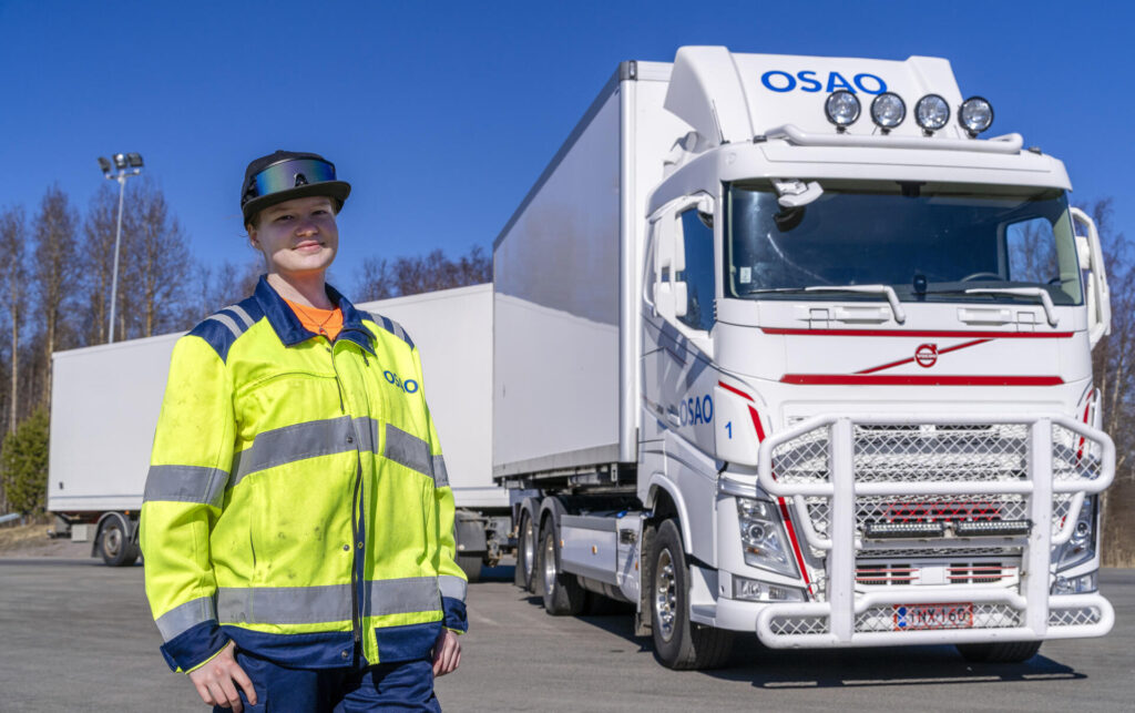 Henkilö heijastavassa takissa seisoo hymyillen yhdistelmäajoneuvon edessä. Kuorma-autossa on teksti 'OSAO'. Taustalla näkyy puita ja kirkas sininen taivas.