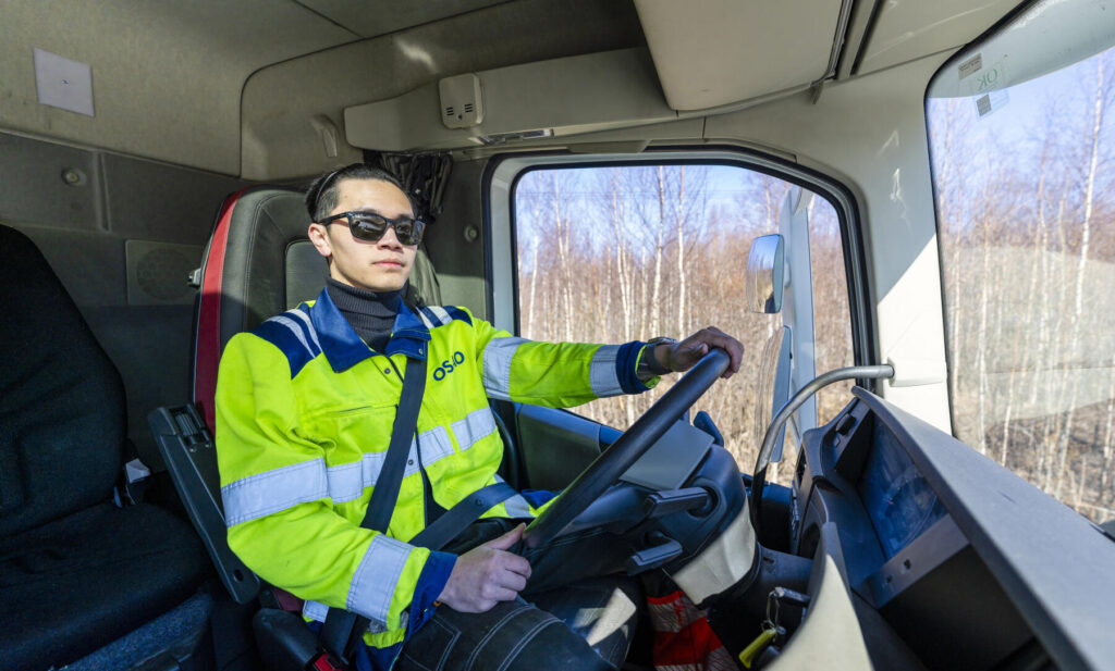 Mies aurinkolaseissa ajaa kuorma-autoa. Hänellä on yllään keltainen huomiotakki ja turvavyö on kiinnitetty.