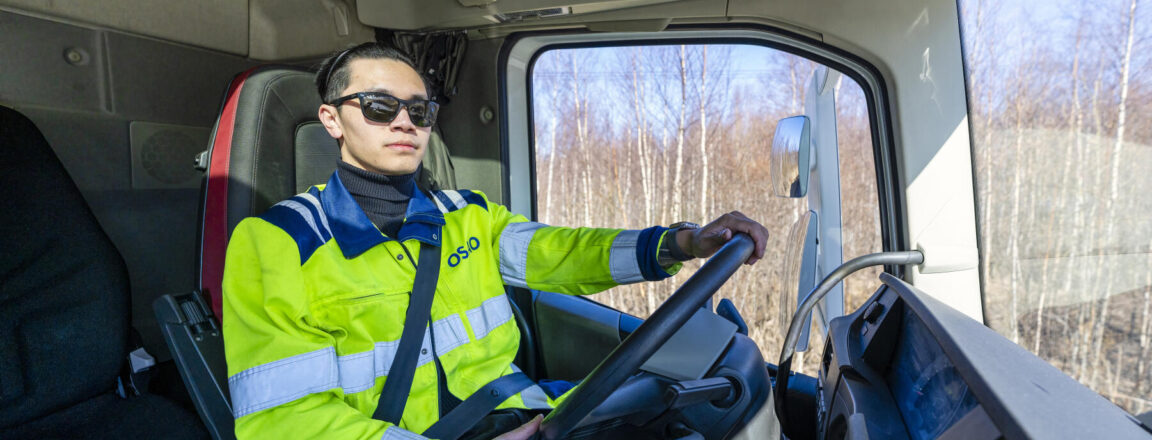 Mies aurinkolaseissa ajaa kuorma-autoa. Hänellä on yllään keltainen huomiotakki ja turvavyö on kiinnitetty.