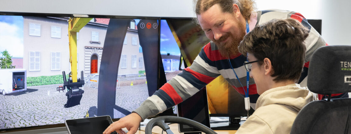 Kuvassa nuori henkilö istuu simulaattorituolissa ja käyttää ajosimulaattoria, jossa on suuri näyttö. Vieressä seisoo opettaja, joka osoittaa simulaattorin näyttöä ja hymyilee.