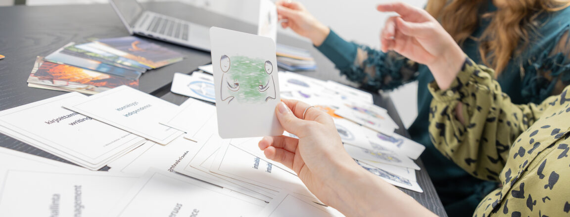 Kaksi henkilöä istuu pöydän ääressä, jolla on paljon kortteja ja kannettava tietokone. Etualalla oleva henkilö pitää kädessään korttia, jossa on piirros kahdesta ihmishahmosta vihreällä taustalla. Henkilöt näyttävät keskustelevan tai valitsevan kortteja.