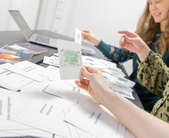 Kaksi henkilöä istuu pöydän ääressä, jolla on paljon kortteja ja kannettava tietokone. Etualalla oleva henkilö pitää kädessään korttia, jossa on piirros kahdesta ihmishahmosta vihreällä taustalla. Henkilöt näyttävät keskustelevan tai valitsevan kortteja.