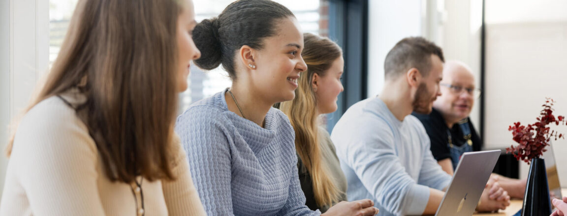 Ryhmä ihmisiä istuu pöydän ääressä keskustelemassa. Heillä on edessään kannettava tietokone ja pöydällä on maljakko, jossa on punaisia koristeita. Taustalla on suuri ikkuna, josta näkyy päivänvalo.