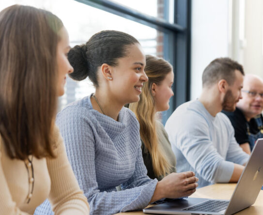 Ryhmä ihmisiä istuu pöydän ääressä keskustelemassa. Heillä on edessään kannettava tietokone ja pöydällä on maljakko, jossa on punaisia koristeita. Taustalla on suuri ikkuna, josta näkyy päivänvalo.