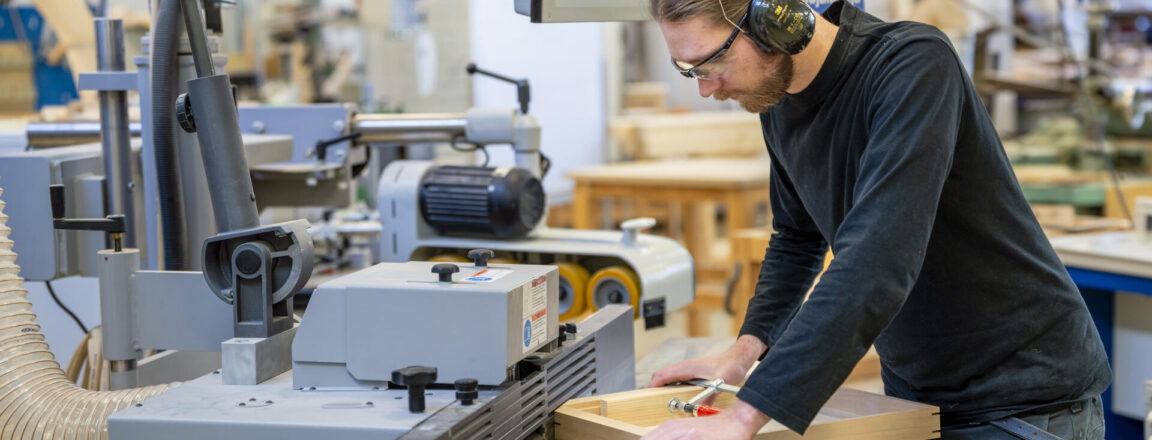 Puuseppä työskentelee puuntyöstökoneen kanssa puusepänverstaalla. Hänellä on kuulosuojaimet ja suojalasit päässään.
