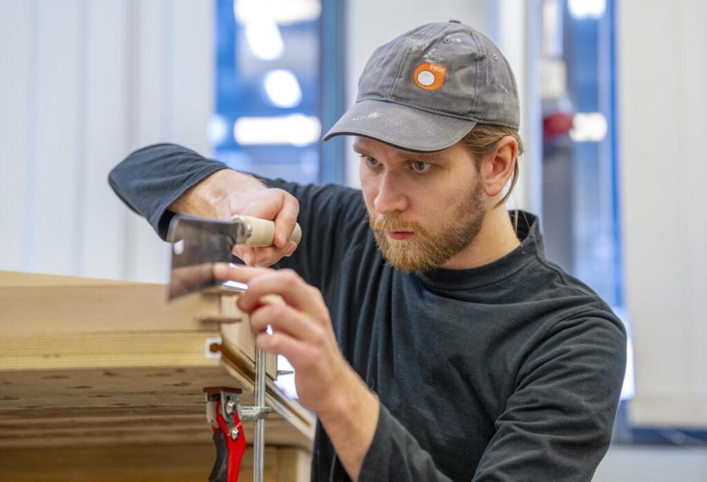 Puuseppä työskentelee keskittyneesti puutyöpajassa. Hän käyttää pientä käsisahaa puupalan leikkaamiseen, ja työpöytä on kiinnitetty puristimella. Miehellä on päässään lippalakki ja yllään musta paita.