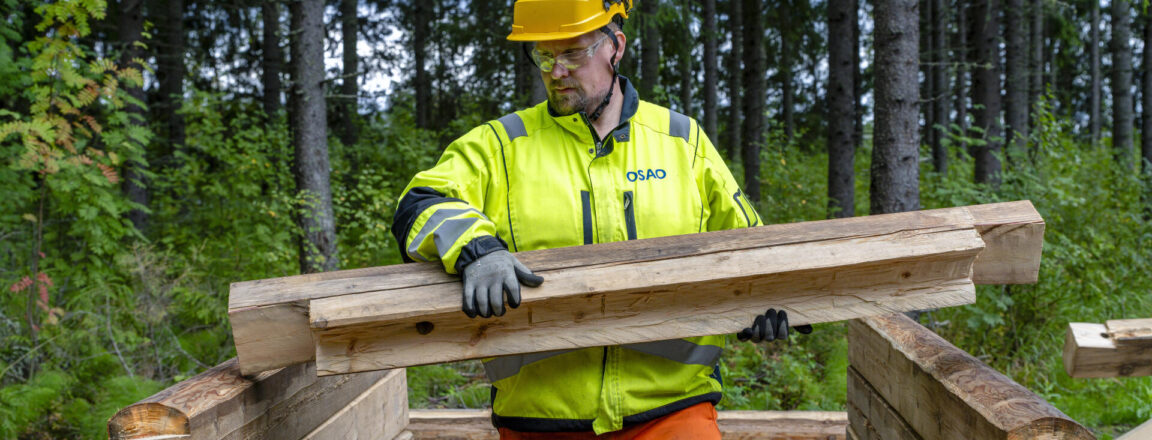 Mies keltaisessa työvaatteessa ja kypärässä rakentaa puista hirsikehikkoa metsässä.