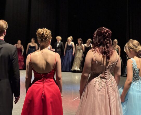 Ryhmän nuoria aikuisia juhlapuvuissa seisoo lavalla odottamassa esityksen alkua.