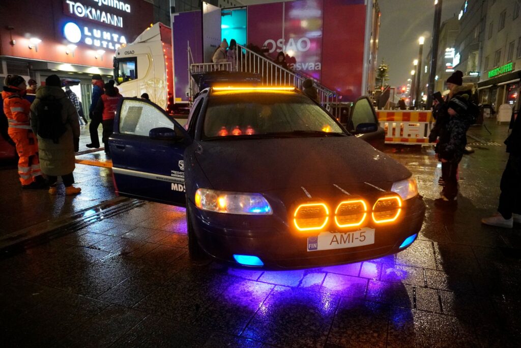Auto, jossa on kirkkaat oranssit ja siniset valot etuosassa, pysäköitynä kaupungin kadulle iltavalaistuksessa. Taustalla on ihmisiä ja liikennettä.