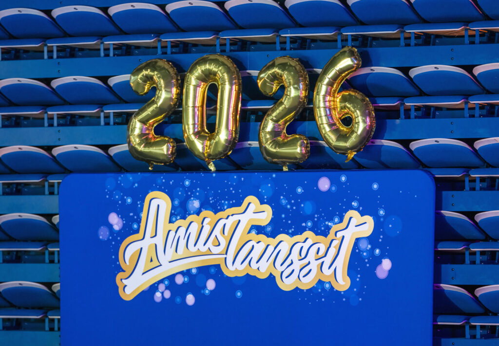 Kuva, jossa on kultaiset ilmapallot, jotka muodostavat numeron 2026, ja alla sininen kyltti, jossa lukee "Amistanssit" valkoisella ja keltaisella tekstillä.