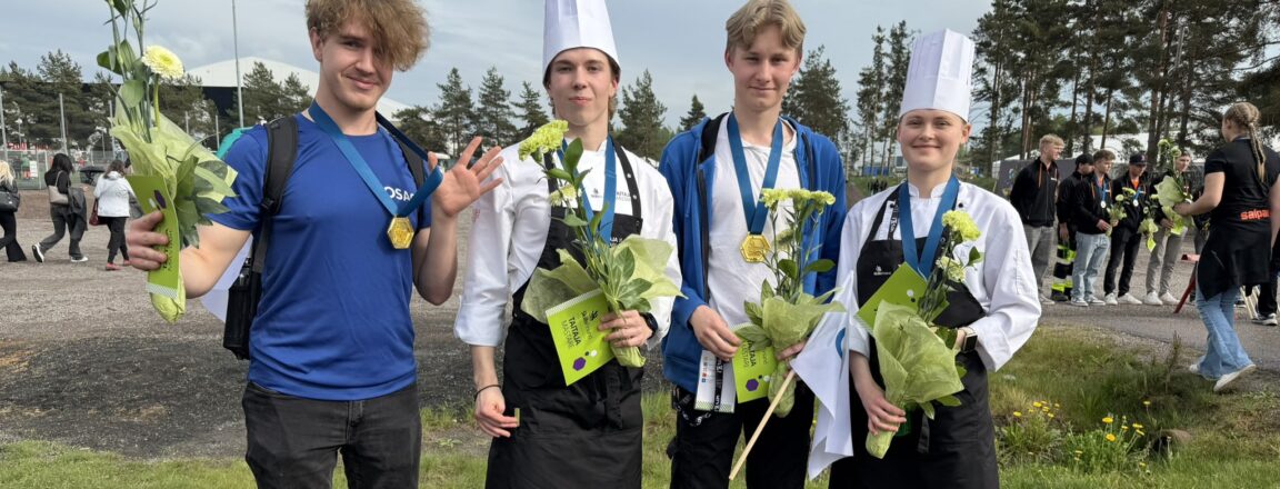 Neljä nuorta henkilöä seisoo ulkona nurmikolla, joista kaksi pukeutunut kokin hattuun ja essuun. Heillä on keltaisia kukkia ja mitalit kaulassa. Taustalla on muita ihmisiä ja puita.