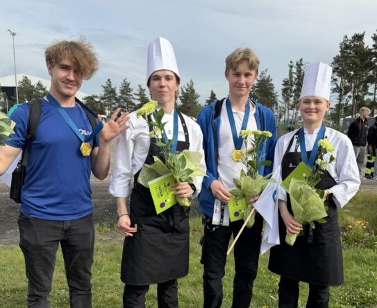 Neljä nuorta henkilöä seisoo ulkona nurmikolla, joista kaksi pukeutunut kokin hattuun ja essuun. Heillä on keltaisia kukkia ja mitalit kaulassa. Taustalla on muita ihmisiä ja puita.