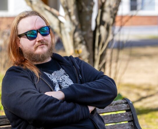Henkilö istuu puistonpenkillä aurinkoisena päivänä. Hänellä on pitkät hiukset, aurinkolasit ja musta huppari. Taustalla näkyy puita ja rakennuksia.