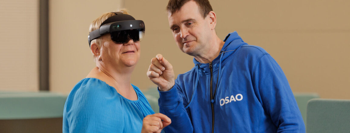 Nainen käyttää virtuaalitodellisuuslaseja, ja mies ohjaa häntä. Miehellä on päällään sininen huppari, jossa lukee 'OSAO'. He ovat sisätiloissa.
