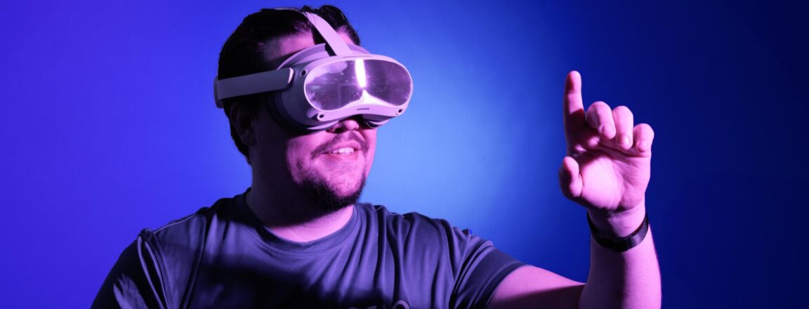 Henkilö käyttää virtuaalitodellisuuslaseja ja osoittaa eteenpäin sormellaan. Tausta on sininen ja valaistus on violetinsävyinen.
