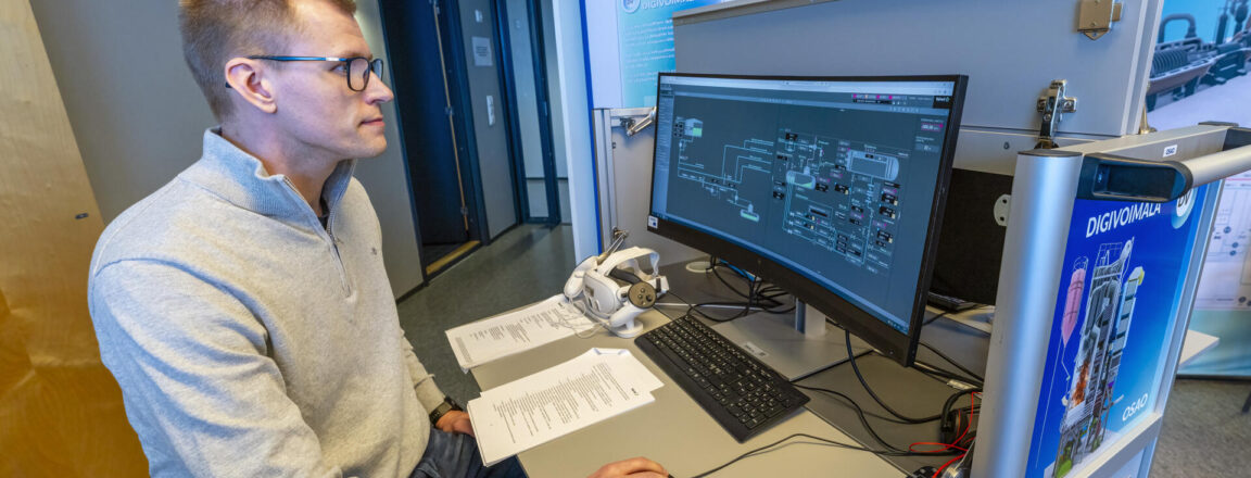 Henkilö työskentelee tietokoneen ääressä digivoimalaan liittyvässä työtilassa. Hän katsoo tietokoneen näyttöä, jossa on kaavioita. Pöydällä on myös papereita ja pienen laitteen osa. Taustalla on sininen mainoskyltti, jossa lukee 'DIGIVOIMALA'.