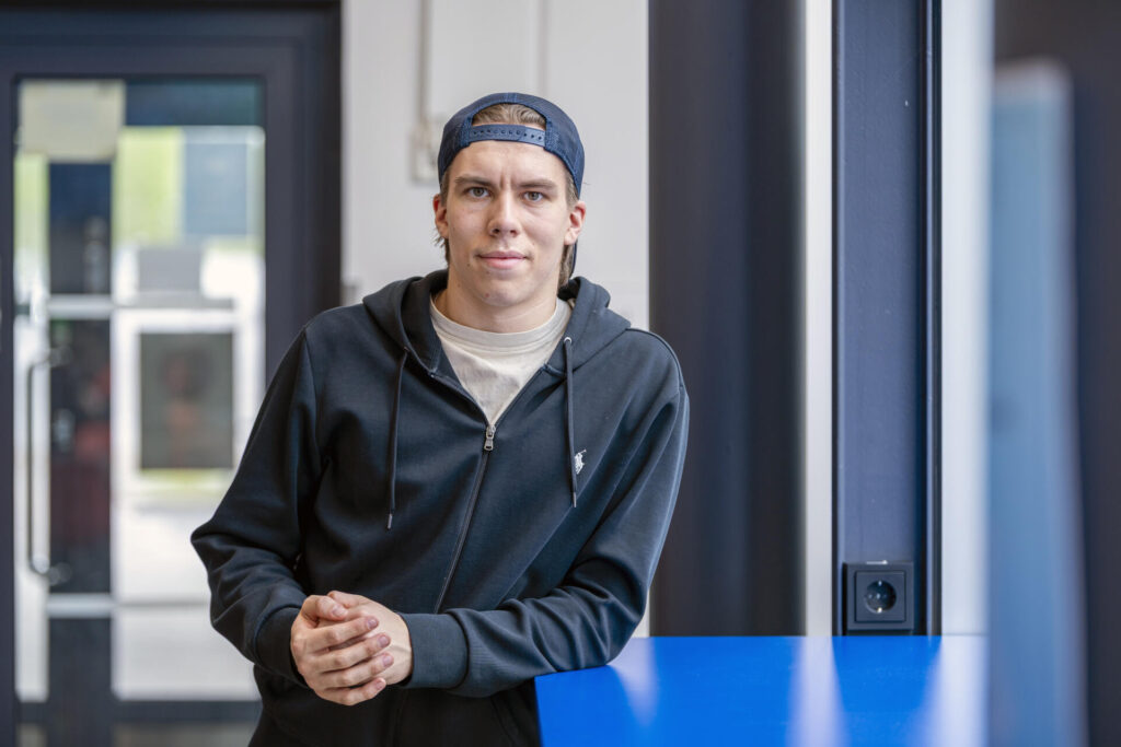 Nuori mies seisoo sisätiloissa päällään musta huppari ja lippalakki. Hän nojaa siniseen pöytään ja katsoo suoraan kameraan.