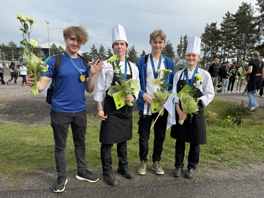 Neljä nuorta seisoo ulkona, kaikilla on kukka kimppu ja mitali kaulassa. Kaksi heistä on pukeutunut kokin takkiin ja hattuun.