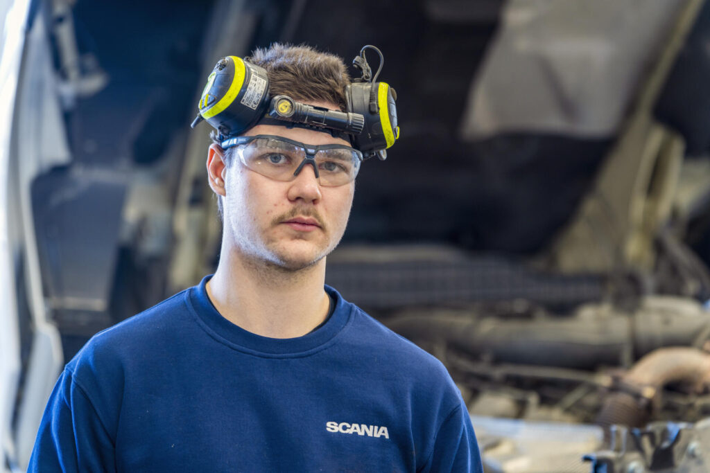 Mies seisoo autokorjaamolla, päässään suojalasit ja kuulosuojaimet. Hänellä on yllään sininen Scania-paita, ja taustalla näkyy kuorma-auton moottori.