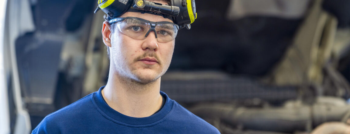 Mies seisoo autokorjaamolla, päässään suojalasit ja kuulosuojaimet. Hänellä on yllään sininen Scania-paita, ja taustalla näkyy kuorma-auton moottori.