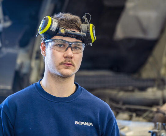 Mies seisoo autokorjaamolla, päässään suojalasit ja kuulosuojaimet. Hänellä on yllään sininen Scania-paita, ja taustalla näkyy kuorma-auton moottori.