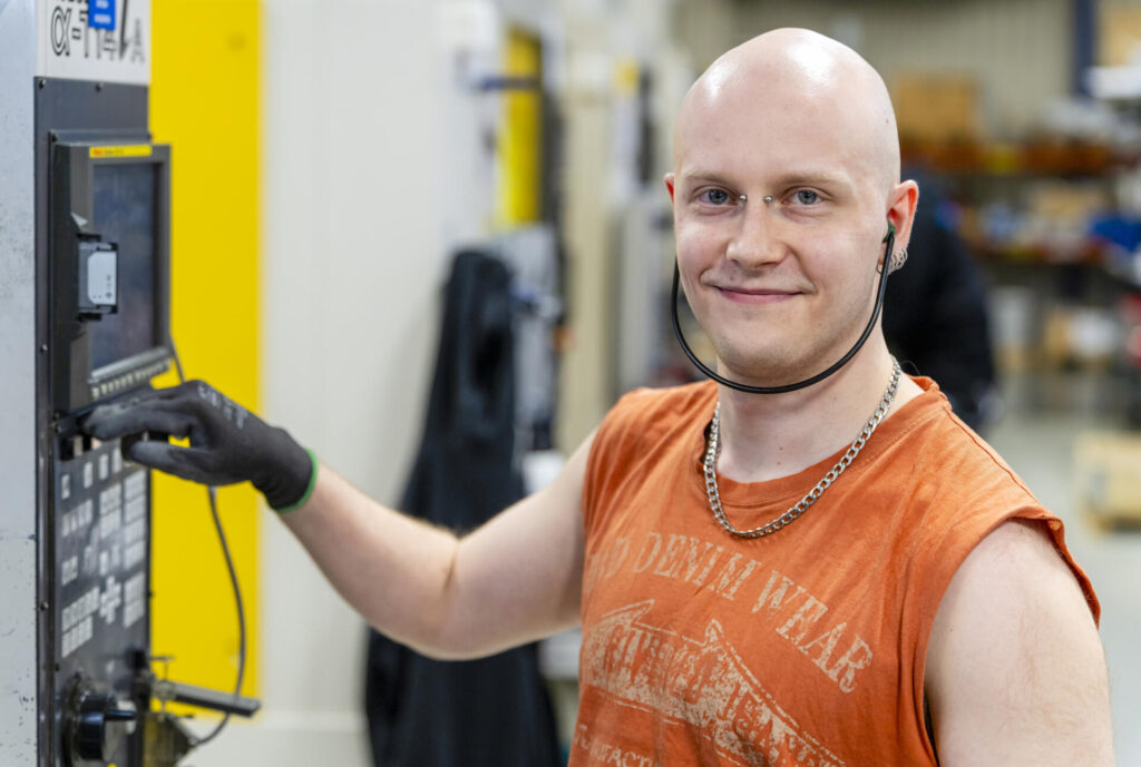 Mies seisoo hymyillen teollisuuslaitteiston edessä ja käyttää sen hallintapaneelia. Hänellä on yllään oranssi hihaton paita ja kuulosuojaimet kaulalla.