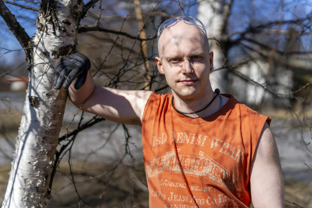 Mies seisoo ulkona aurinkoisena päivänä, pukeutuneena oranssiin hihattomaan paitaan ja suojalaseihin, nojaten koivunrunkoon.
