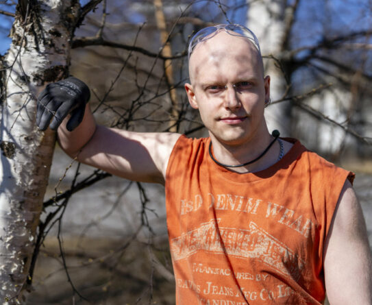 Mies seisoo ulkona aurinkoisena päivänä, pukeutuneena oranssiin hihattomaan paitaan ja suojalaseihin, nojaten koivunrunkoon.
