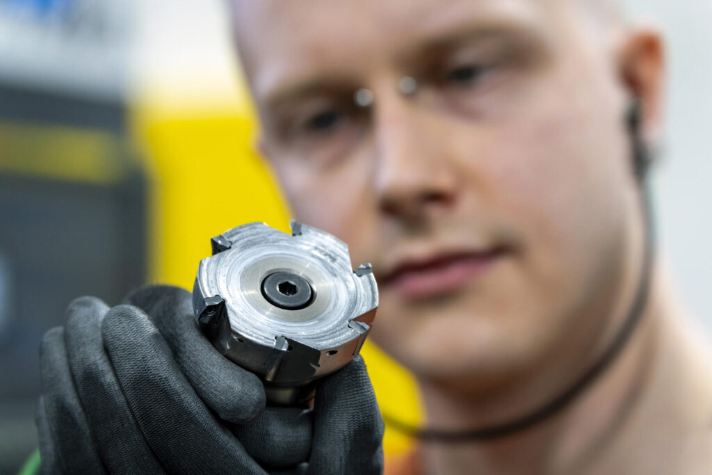 Henkilö pitää käsissään metallista kappaletta teollisuusympäristössä. Hänen katseensa on keskittynyt kädessään olevaan osaan, ja hänellä on käsineet kädessään ja kuulosuojaimet korvilla.