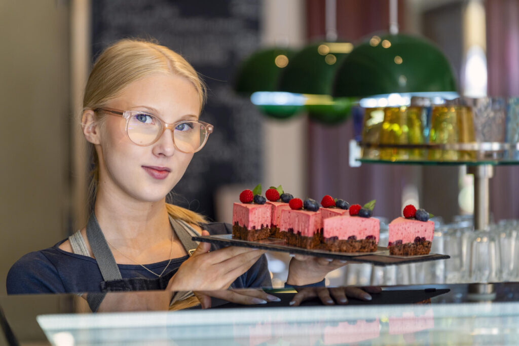 Nainen esittelee tarjotinta, jolla on vaaleanpunaisia kakkupaloja koristeltuna marjoilla. Hänellä on päällään essu ja silmälasit. Taustalla näkyy vihreitä valaisimia ja hylly.