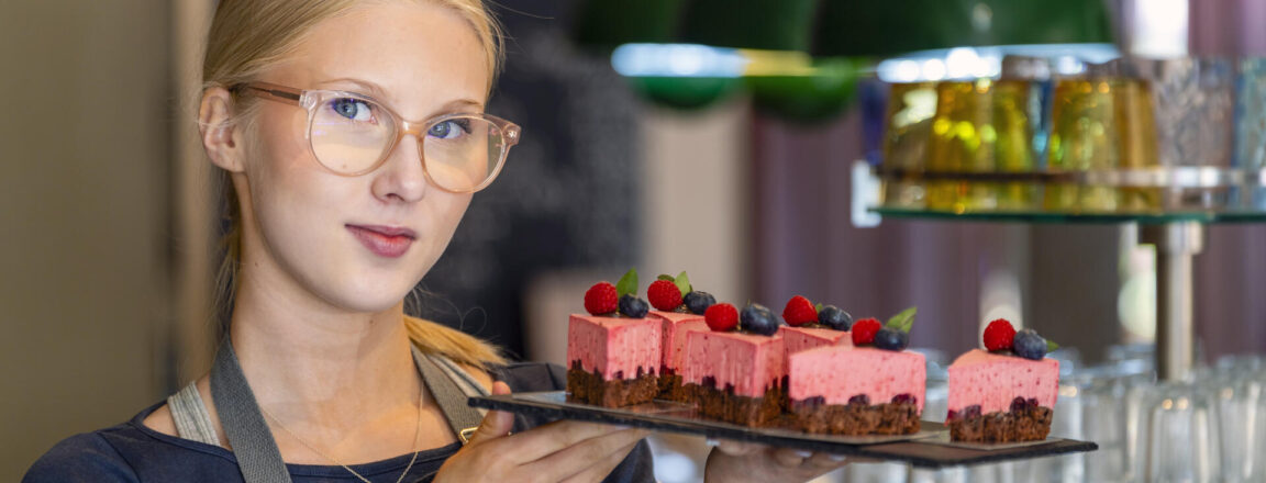Nainen esittelee tarjotinta, jolla on vaaleanpunaisia kakkupaloja koristeltuna marjoilla. Hänellä on päällään essu ja silmälasit. Taustalla näkyy vihreitä valaisimia ja hylly.