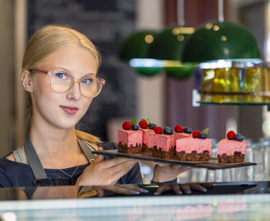 Nainen esittelee tarjotinta, jolla on vaaleanpunaisia kakkupaloja koristeltuna marjoilla. Hänellä on päällään essu ja silmälasit. Taustalla näkyy vihreitä valaisimia ja hylly.