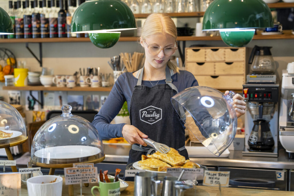 Nainen asettelee leivonnaisia esille kahvilassa. Naisen on kahvilan työntekijä ja hänellä on päällään Paulig-esiliinaa.