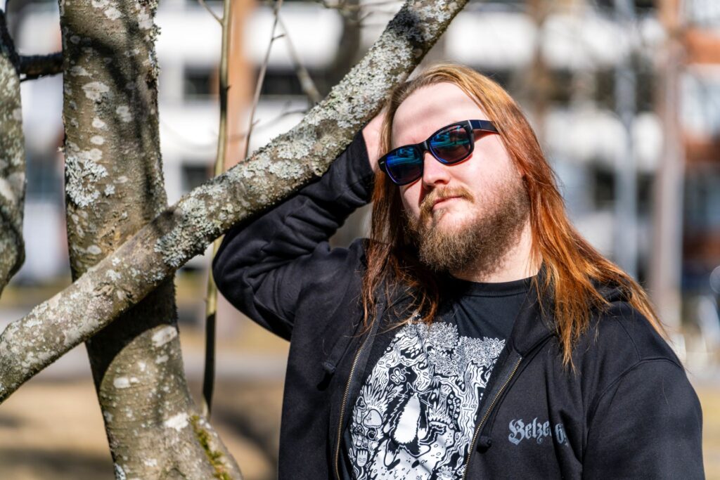 Henkilö seisoo nojaamassa puun oksaan aurinkoisena päivänä. Hänellä on aurinkolasit, pitkät punaruskeat hiukset, parta ja musta huppari, jossa on koristeellinen kuvio. Taustalla näkyy sumeasti rakennuksia.