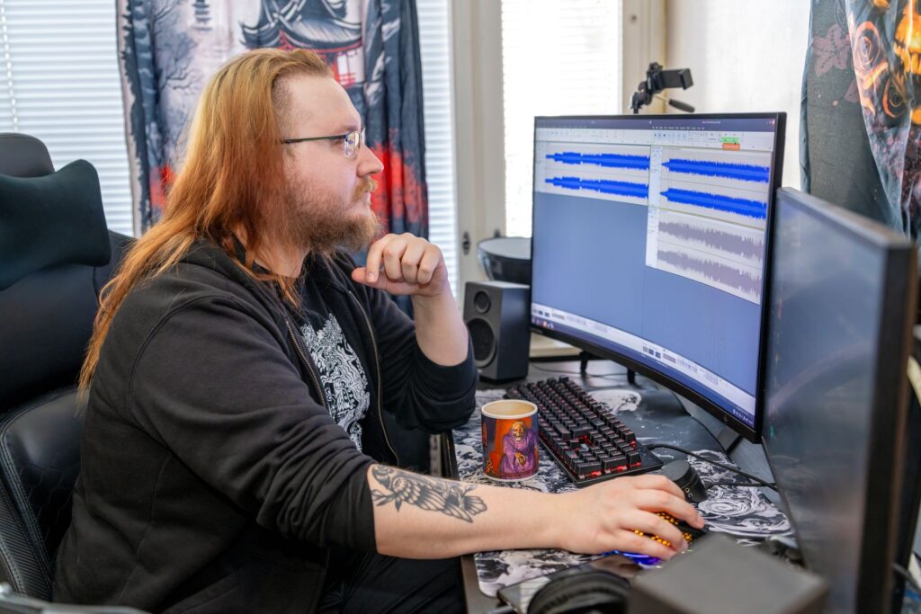Henkilö työskentelee tietokoneella, jossa on auki äänieditoriohjelma. Hänellä on punaiset hiukset ja silmälasit. Pöydällä on kahvikuppi ja näppäimistö.