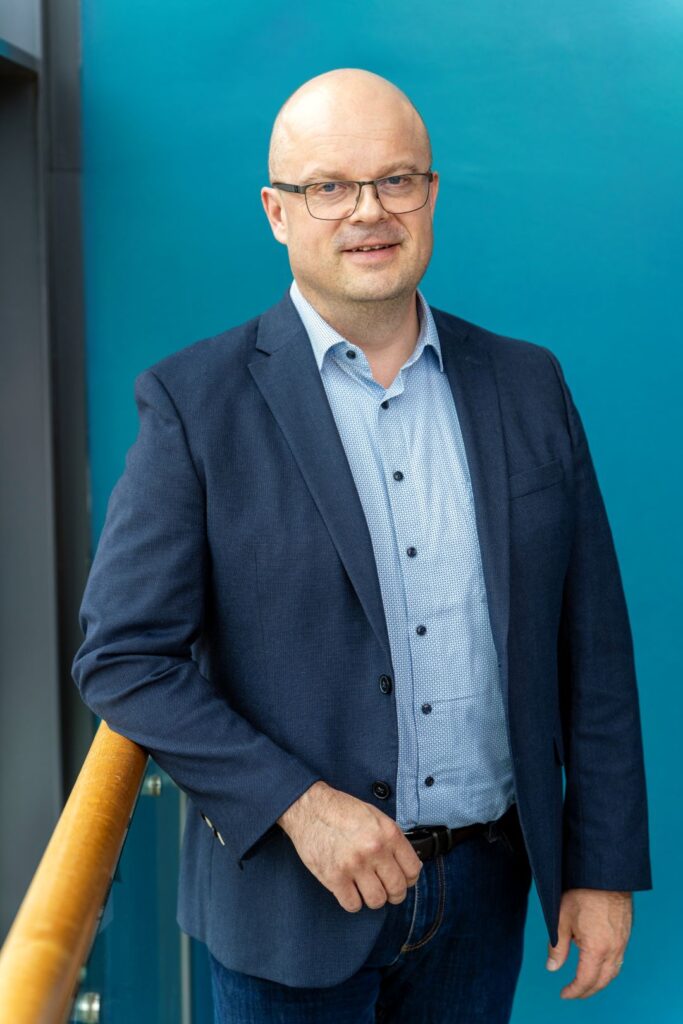 Mies seisoo sinistä seinää vasten pukeutuneena siniseen pukuun ja vaaleansiniseen kauluspaitaan, nojaten kaiteeseen.