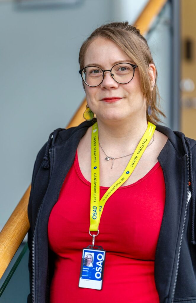 Henkilö seisoo portaiden vieressä hymyillen. Hänellä on silmälasit, punainen paita ja musta huppari. Kaulassa roikkuu keltainen henkilökorttinauha, jossa lukee 'Kulttuuriagentti'. Henkilökortissa on sininen tausta ja OSAO-logo.