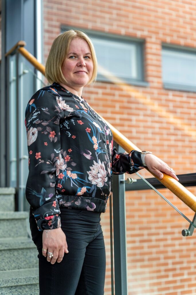 Nainen seisoo portaiden vieressä pitäen kaiteesta kiinni. Hänellä on kukkakuvioinen pusero ja taustalla näkyy tiiliseinä.