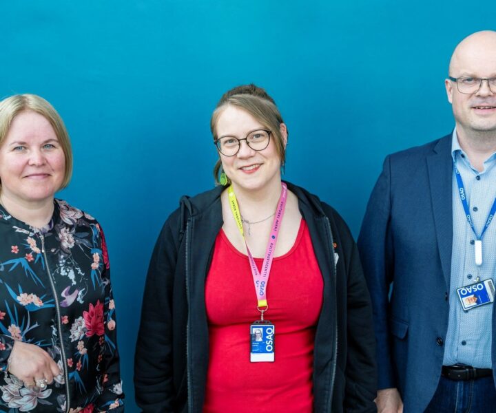Kolme henkilöä seisoo rinnakkain. Vasemmalla henkilö, jolla on vaalea tukka ja kukkakuviollinen paita. Keskellä henkilö, jolla on pitkät ruskeat hiukset, silmälasit ja punainen paita. Oikealla henkilö, jolla on kalju pää, silmälasit ja sininen puku. Kaikilla on kaulassa nimikortit.