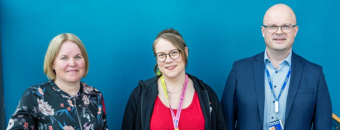 Kolme henkilöä seisoo rinnakkain. Vasemmalla henkilö, jolla on vaalea tukka ja kukkakuviollinen paita. Keskellä henkilö, jolla on pitkät ruskeat hiukset, silmälasit ja punainen paita. Oikealla henkilö, jolla on kalju pää, silmälasit ja sininen puku. Kaikilla on kaulassa nimikortit.