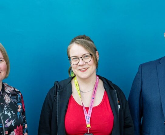 Kolme henkilöä seisoo rinnakkain. Vasemmalla henkilö, jolla on vaalea tukka ja kukkakuviollinen paita. Keskellä henkilö, jolla on pitkät ruskeat hiukset, silmälasit ja punainen paita. Oikealla henkilö, jolla on kalju pää, silmälasit ja sininen puku. Kaikilla on kaulassa nimikortit.