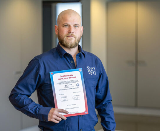 Henkilö seisoo sisätiloissa pitäen kädessään International Institute of Welding -todistusta. Hänellä on päällään sininen kauluspaita, jossa on Syri & Syri -teksti. Taustalla on vaaleat seinät ja suljetut ovet.