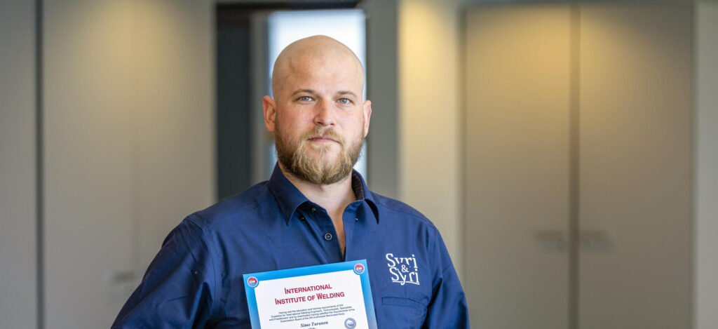Henkilö sinisessä kauluspaidassa pitää kädessään todistuksen, jossa lukee 'International Institute of Welding'. Taustalla näkyy vaalea huone.