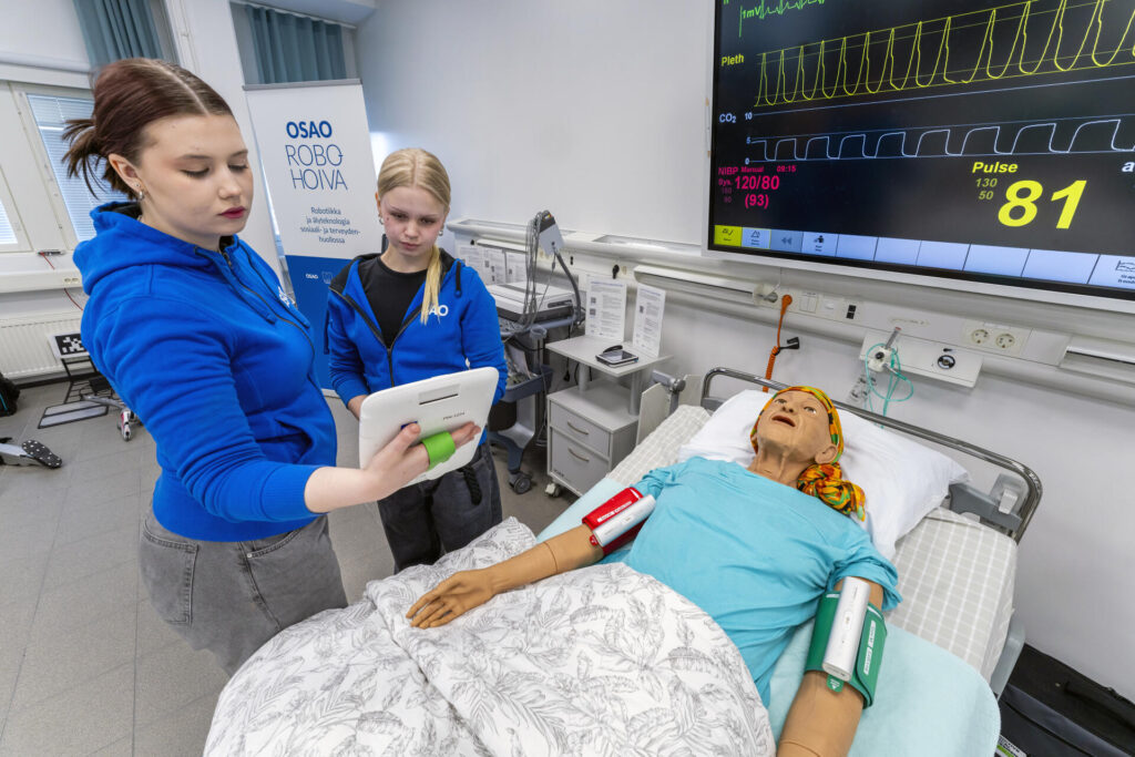 Kaksi opiskelijaa sinisissä huppareissa tarkastelevat tablettia sairaalaympäristössä, jossa on vuoteella makaava simulaationukke. Seinällä näkyy näyttö, jossa on elintoimintoja kuvaavia tietoja, kuten pulssi ja verenpaine.